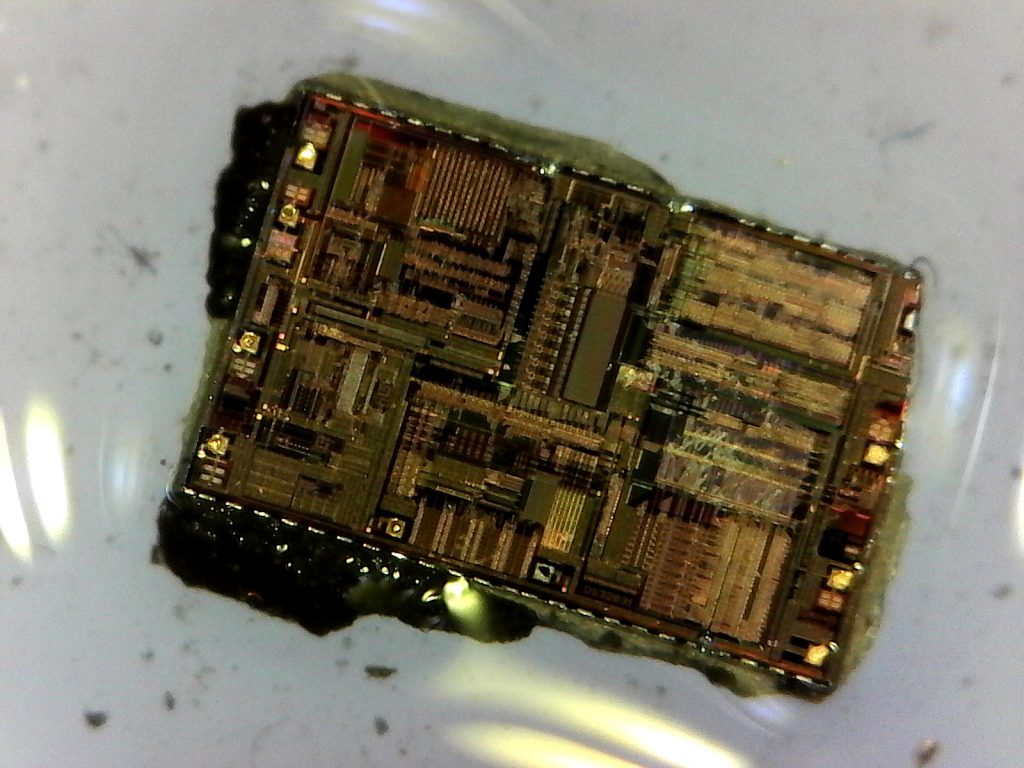 The whole DS3231 die. The die is still wet with isopropanol and bits of the epoxy package are still stuck to the outer edges of the die. Scratches appear on the several parts of the complex circuit laid out on the die.